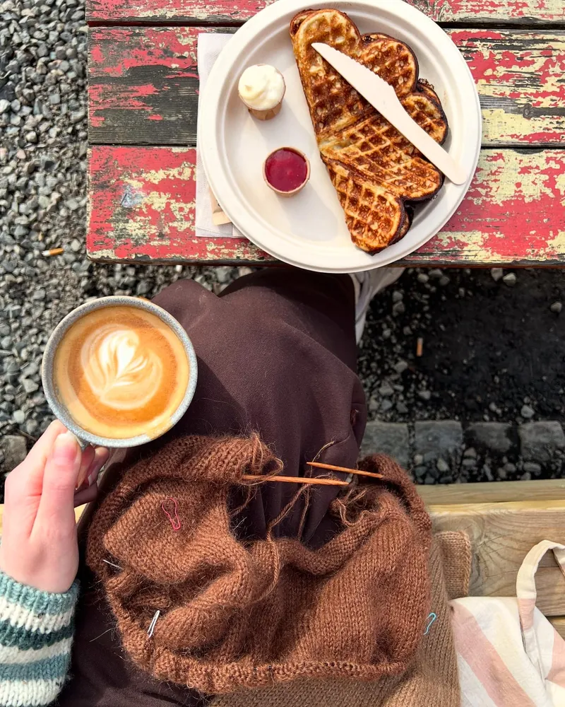 Kaffe med hjerte i melkeskummet, vafler og et brunt strikketøy på fanget ved et kafébord ute