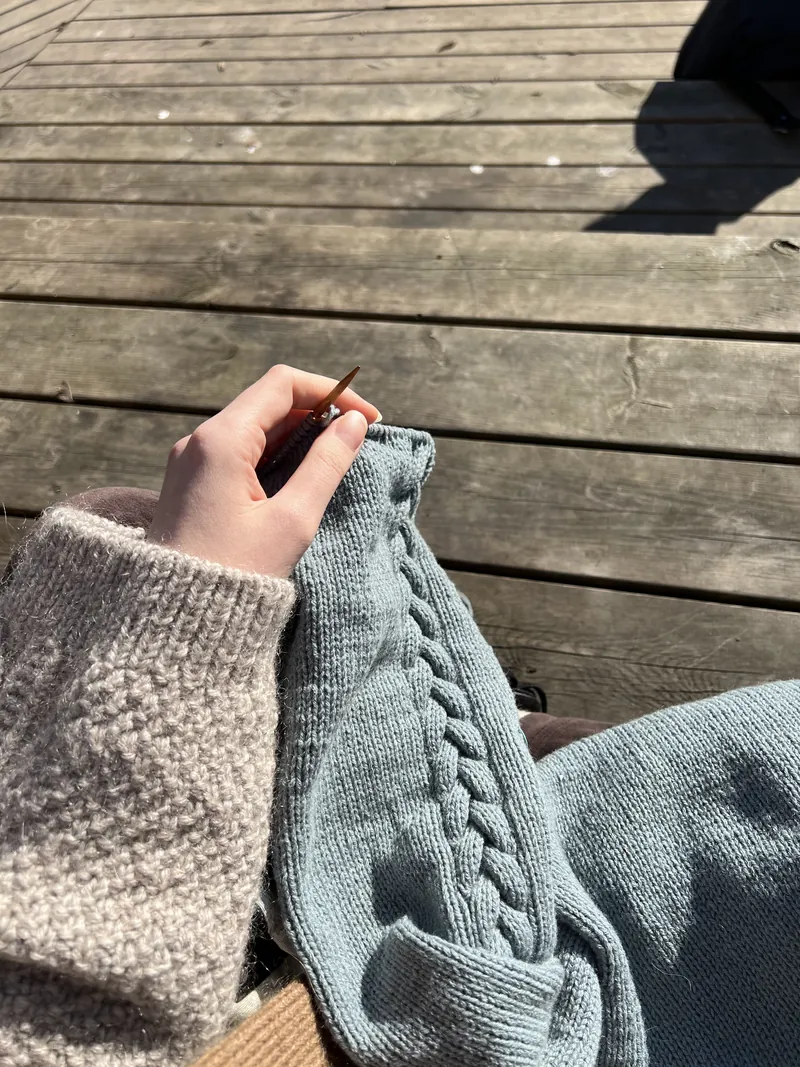 Lyseblått erme med flette som strikkes i sola på en treterrasse