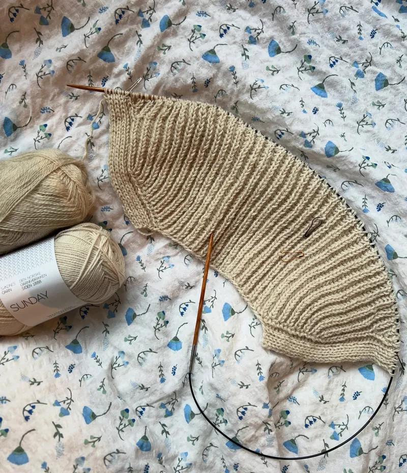 Beige ribbestrikk på rundpinne og to garnnøster på en blomstrete dyne
