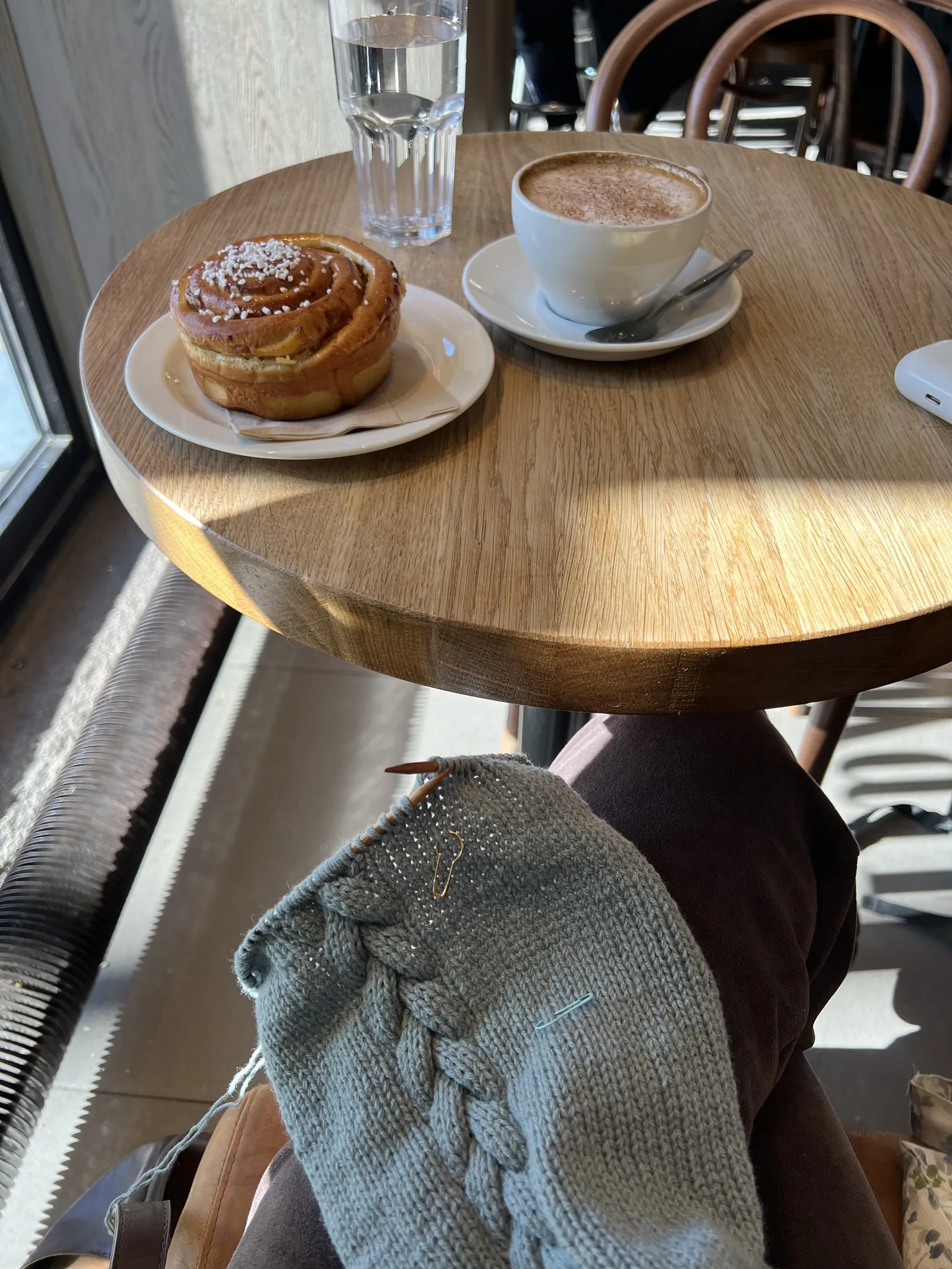 Kafébord ved vinduet med kaffe og kanelbolle, og et lyseblått strikket erme med flette